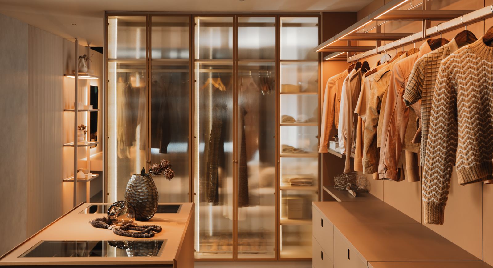 Closet aberto com portas de vidro iluminadas, araras de roupas e bancada em Favo e Teron.
